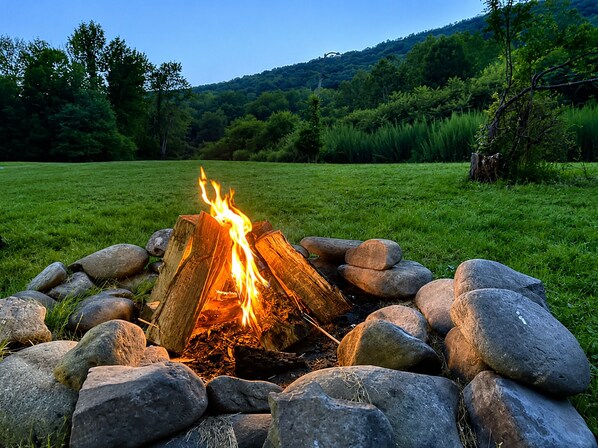 A campfire surrounded by rocks in a grassy field with trees and hills in the background.