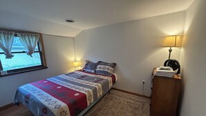 A bedroom with a bed, a window with curtains, a bedside table with a lamp, and a wall socket.