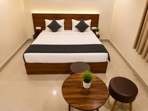 Una habitación de hotel con una cama grande, mesitas de noche, una mesa redonda de madera con una planta y un taburete marrón.