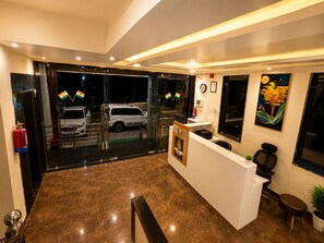 Zona de recepción de un hotel con suelo de mármol, mostrador de recepción, una silla, una mesita, una planta en maceta y un cuadro en la pared.