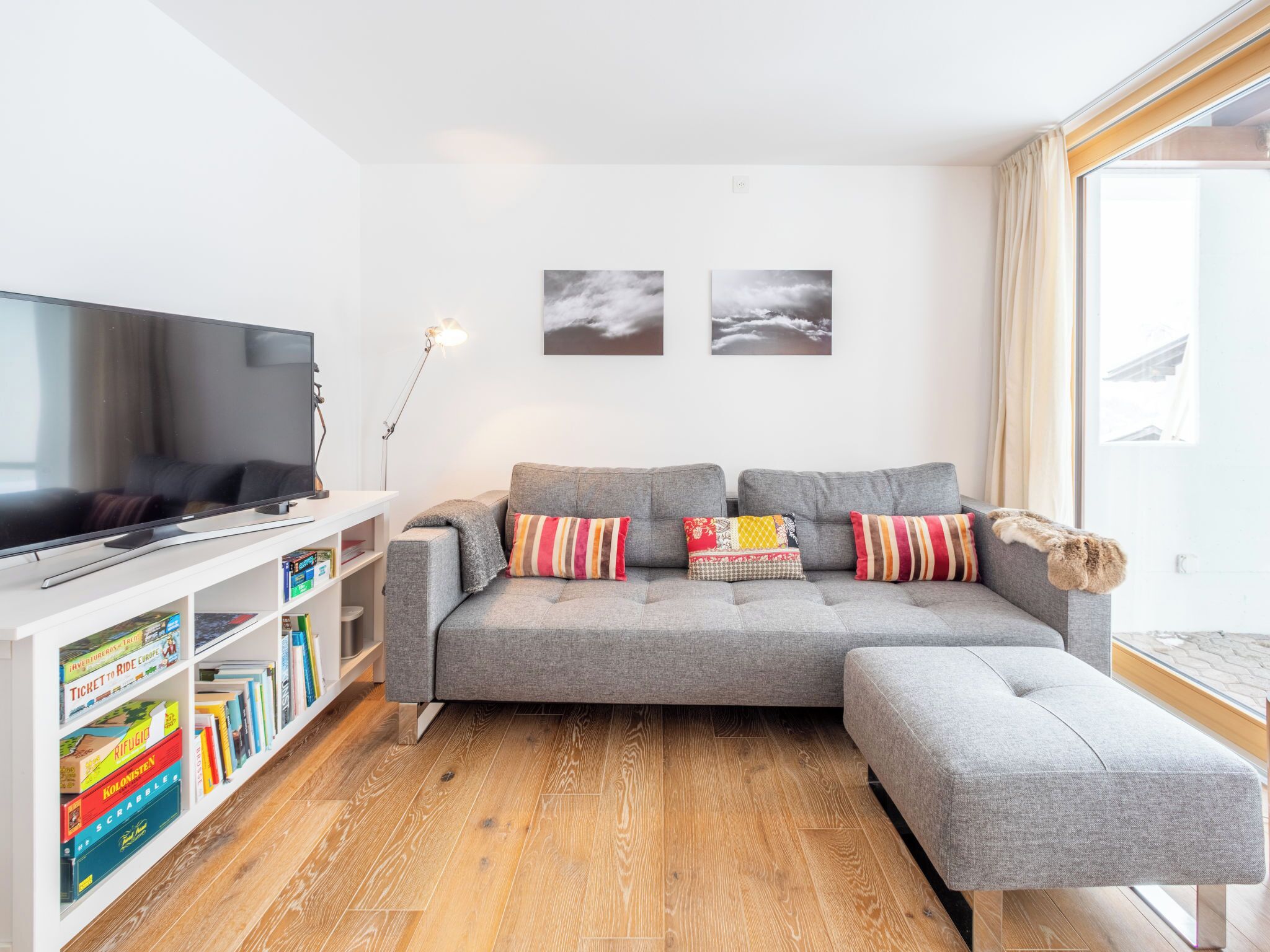 A modern living room with a grey sofa, a flat-screen TV, a bookshelf with travel books, and two framed pictures on the wall.