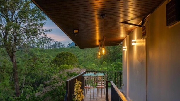 A balcony with a view of a forested landscape, a table and chairs, and a wooden ceiling with hanging lights.