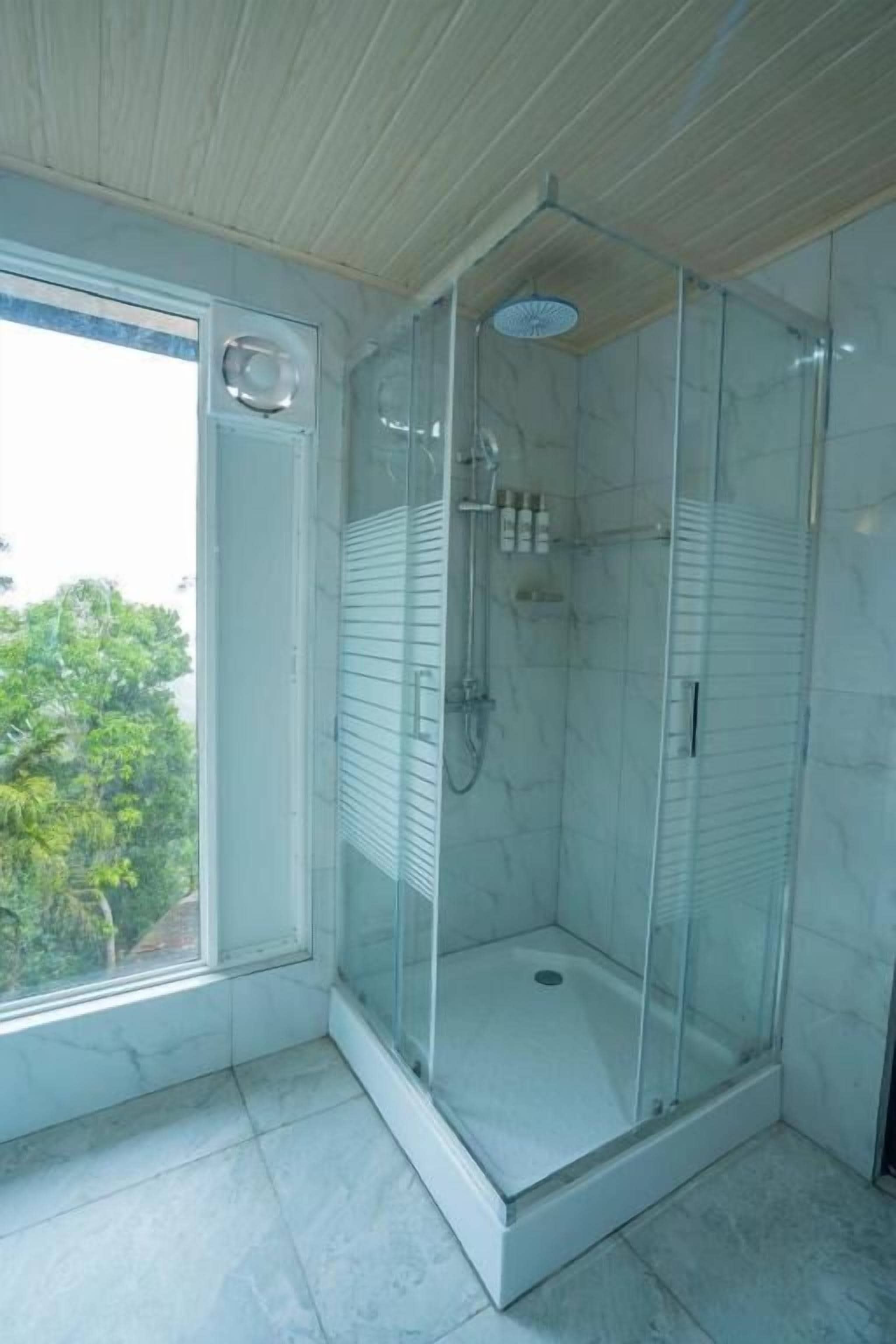 Una ducha con paredes de vidrio, suelo de baldosas blancas y una ventana lateral.