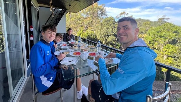 A group of people dining at an outdoor table with a glass top, surrounded by a railing, overlooking a forested landscape.