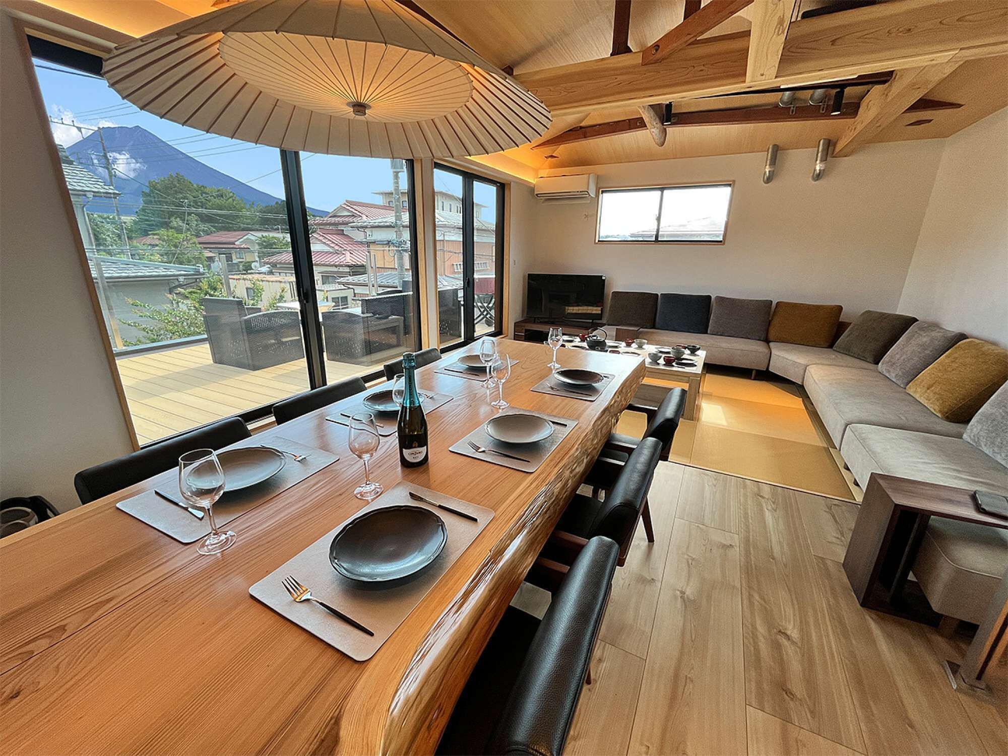 A modern dining area with a long wooden table set for six, large windows offering a view of a mountain, and a cozy seating area with a sofa and chairs.