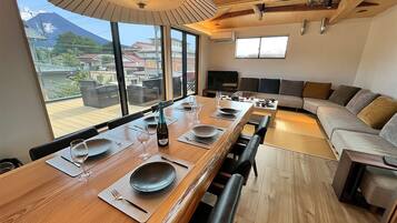 A modern dining area with a long wooden table set for six, large windows offering a view of a mountain, and a cozy seating area with a sofa and chairs.