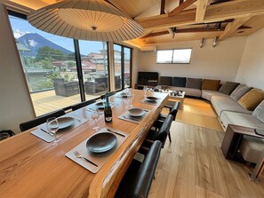 A modern dining area with a long wooden table set for six, large windows offering a view of a mountain, and a cozy seating area with a sofa and chairs.