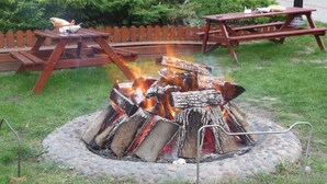 Un foyer avec des bûches, entouré d’une bordure en pierre, et une table de pique-nique en bois à l’arrière-plan.