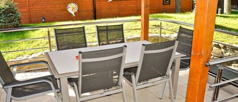 An outdoor seating area with a table and chairs, a wooden building in the background, and a hanging flower basket.