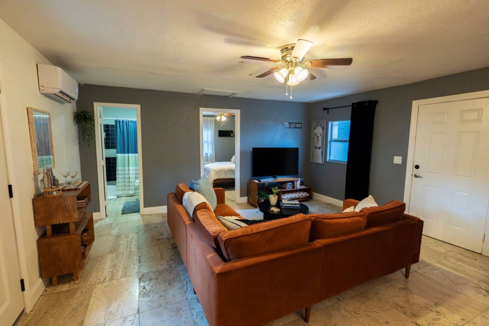 A living room with a brown sofa, a ceiling fan, a flat-screen TV, and a door labeled 'GUEST ROOM'.