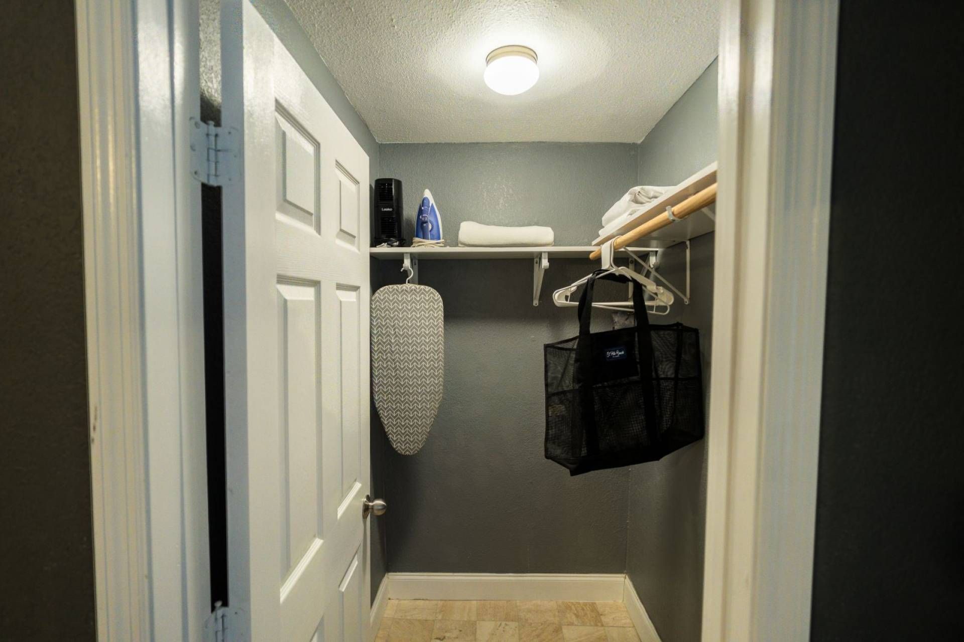 A narrow hallway with a door, a hanging ironing board, and a shelf with various items.
