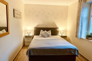 A bedroom with a bed, two bedside tables, a nightstand, a mirror, a window with curtains, and a wall with a floral pattern. - Feuerstein House Mönchgut (Mönchgut)