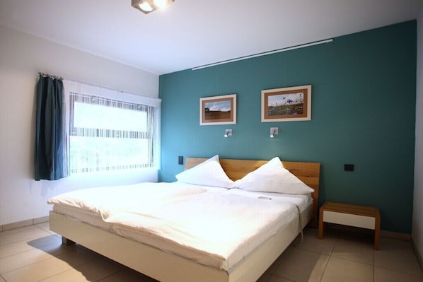 Ein Schlafzimmer mit einem Bett, einem Nachttisch, zwei gerahmten Bildern an der Wand und einem Fenster mit Vorhängen.