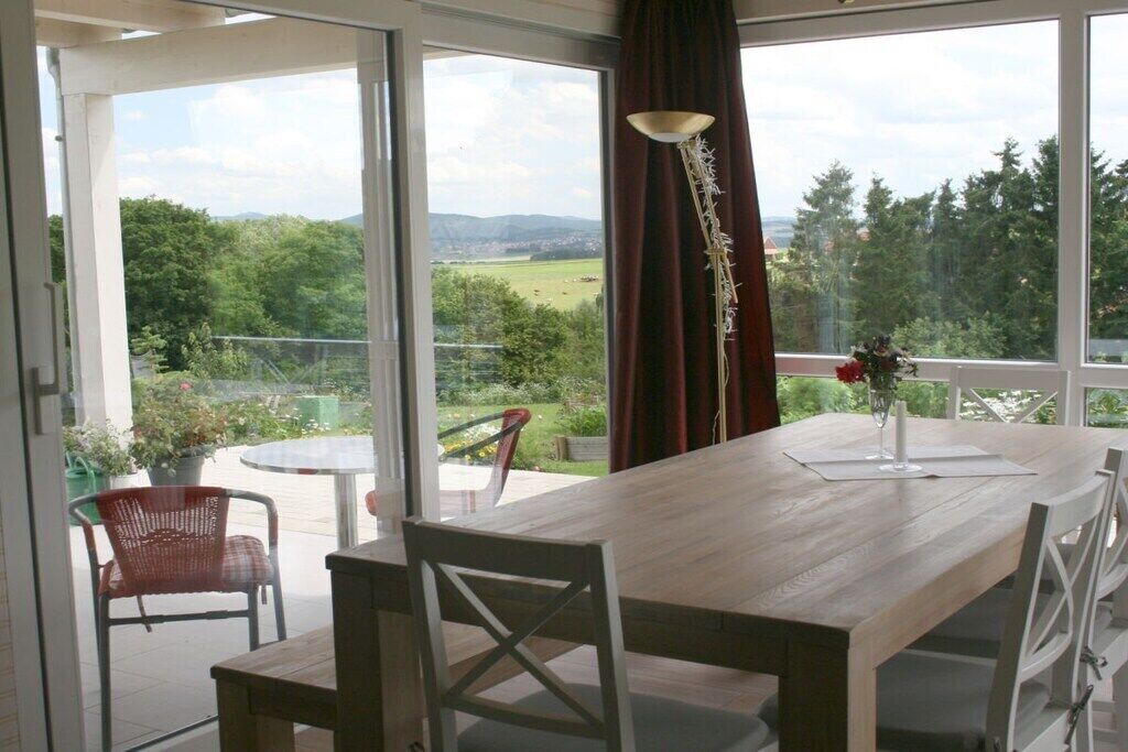 A dining area with a wooden table and chairs, a view of greenery and hills through large windows, and a decorative lamp.
