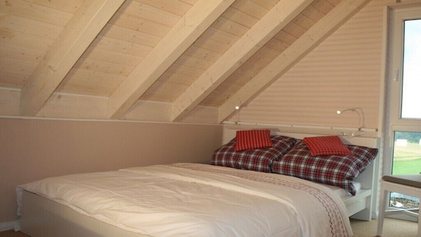 Ein Schlafzimmer mit schrägen Wänden, einem Bett mit weißer Bettwäsche und einem Fenster mit Blick.