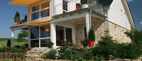A two-story house with a balcony, stone foundation, and a garden with various plants.