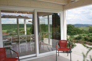 Um alpendre envidraçado com mobiliário de exterior, uma cadeira vermelha e vista para uma paisagem verde.