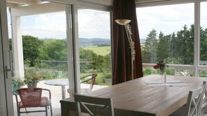 Ein Essbereich mit einem Holztisch und Stühlen, Blick auf Grünflächen und Hügel durch große Fenster sowie eine dekorative Lampe.