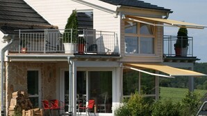 Ein zweistöckiges Haus mit Balkon, Sitzgelegenheit im Freien und Topfpflanzen.