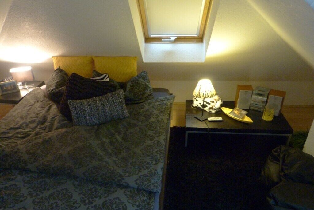 A bedroom with a bed, a desk with a lamp, a small table with papers, and a window with a view.