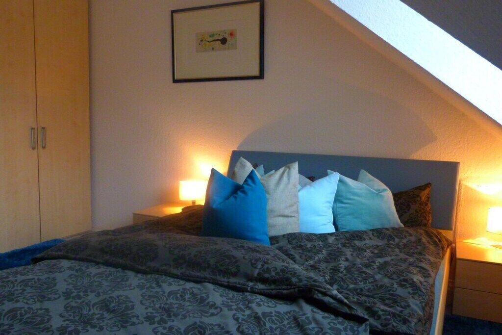 Een slaapkamer met een bed, kussens, een nachtkastje en een ingelijst foto aan de muur.