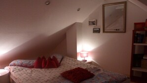 Un dormitorio con cama, almohadas, mesita de noche, lámpara, espejo y estantería.