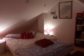 Ein Schlafzimmer mit einem Bett, Kissen, einem Nachttisch, einer Lampe, einem Spiegel und einem Bücherregal.