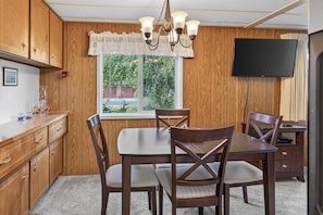 A compact kitchen with wooden cabinets, a dining table with four chairs, a television mounted on the wall, and a window with a view of greenery outside. - Close to Beach & Dtwn! Extended Long Beach Stay (Long Beach)