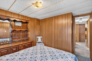 Ein Schlafzimmer mit Holzwandverkleidung, einem Bett mit gemustertem Überwurf, einem Schminktisch mit Spiegel und einer Tür zu einem weiteren Raum.