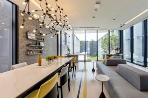 A modern interior with a dining area, a sofa, and a large window offering a view of the outdoors. - Top-Floor Condo Next to MRT Blue Line, Near Malls, Pool, Gym & Quiet Living (Krung Thep Maha Nakhon)