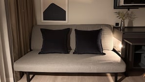 A modern living room with a grey sofa, two black cushions, a framed abstract artwork, a microwave, and a small plant on a side table. - Top-Floor Condo Next to MRT Blue Line, Near Malls, Pool, Gym & Quiet Living (Krung Thep Maha Nakhon)