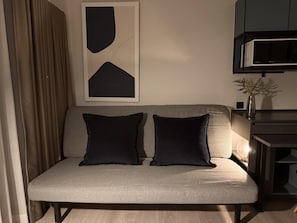 Ein modernes Wohnzimmer mit einem grauen Sofa, zwei schwarzen Kissen, einem gerahmten abstrakten Kunstwerk, einer Mikrowelle und einer kleinen Pflanze auf einem Beistelltisch.