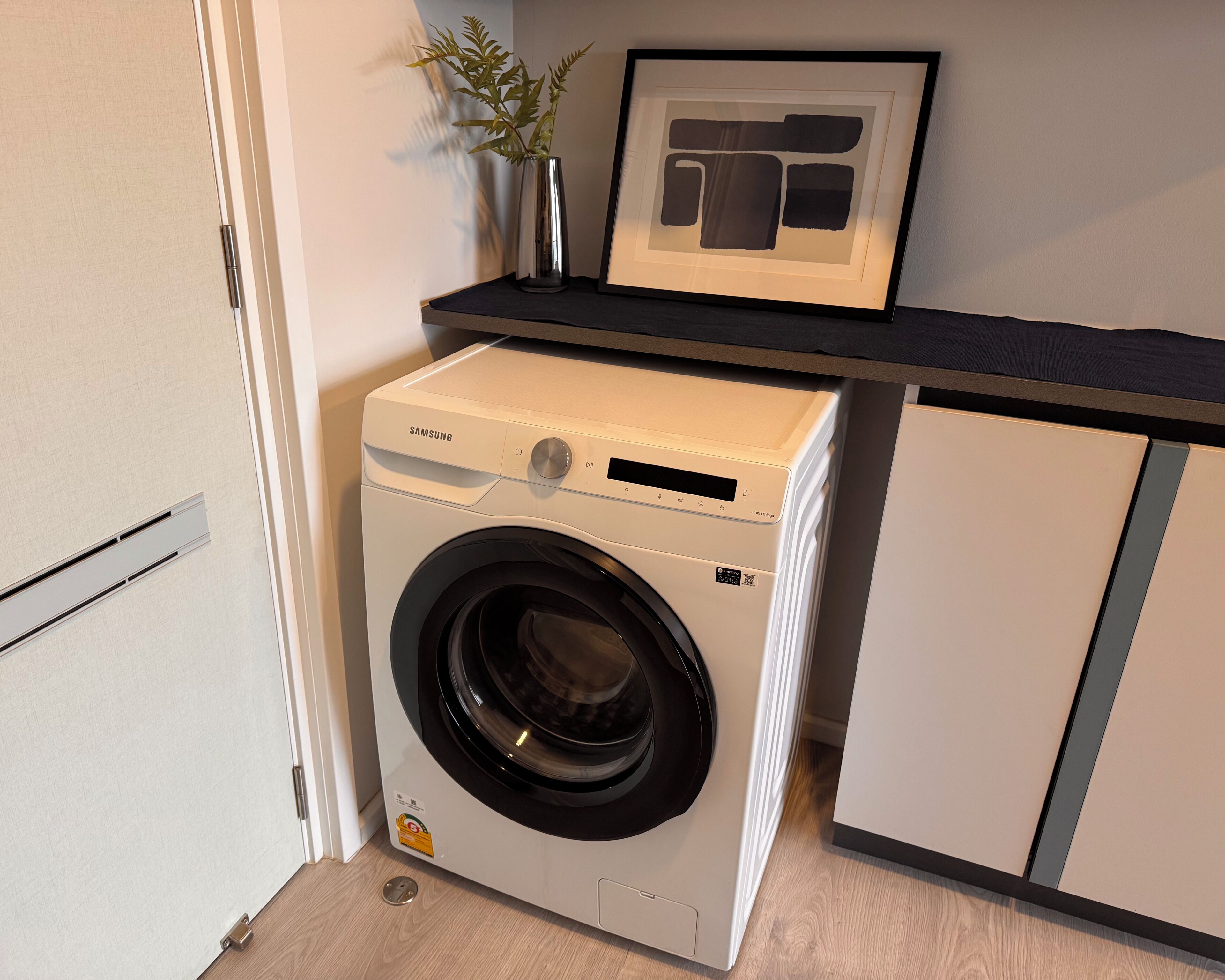 A compact, white Samsung washing machine with a black door, placed next to a white cabinet in a small laundry area.