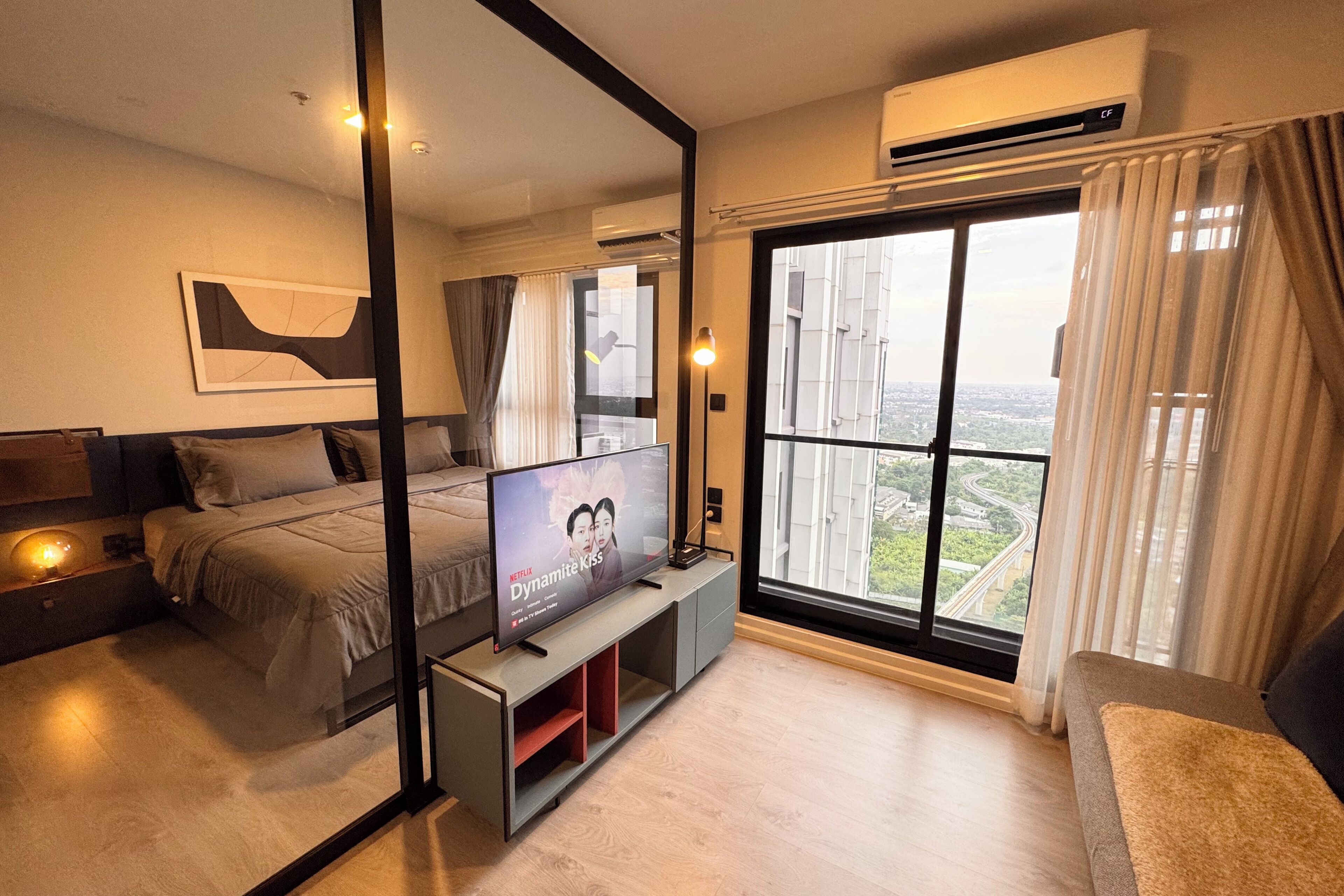 A modern hotel room with a large bed, a TV on a stand, a large window with a view of the city, and an air conditioning unit.