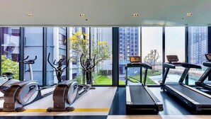A gym with cardio equipment, a treadmill, and a stationary bike, with a view of buildings and greenery. - Top-Floor Condo Next to MRT Blue Line, Near Malls, Pool, Gym & Quiet Living (Krung Thep Maha Nakhon)