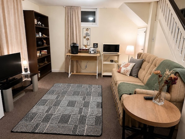 A cozy living room with a sofa, a coffee table, a TV, a bookshelf, and a desk with a computer.
