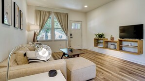 A modern living room with a sofa, ottoman, coffee table, floor lamp, and a flat-screen TV on a wooden shelf. - Historic Southside Fort Worth Home w/ Fenced Yard! (Fort Worth)