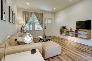 Ein modernes Wohnzimmer mit Sofa, Hocker, Couchtisch, Stehlampe und einem Flachbildfernseher auf einem Holzregal.