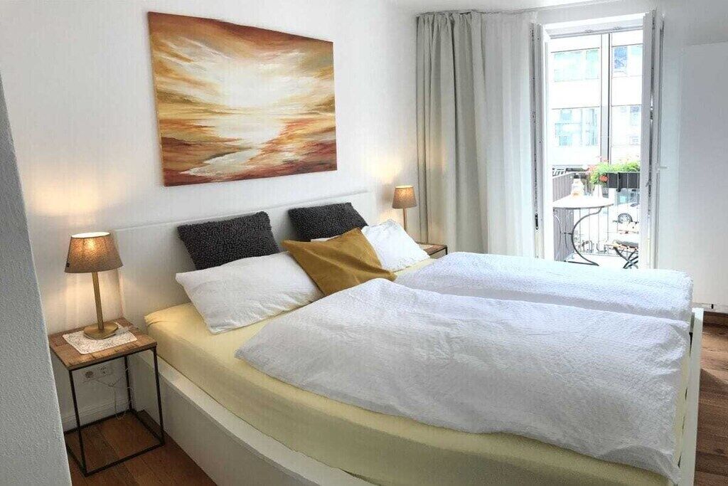 Ein Hotelzimmer mit zwei Betten, einem Bild an der Wand, einem Fenster mit Vorhängen und einem kleinen Tisch mit einer Lampe.