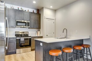 A modern kitchen with stainless steel appliances, a central island with stools, and a white door. - Explore Glen Canyon/Lake Powell! Red River Retreat (Page)