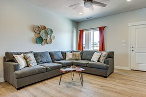 A living room with a sectional sofa, a coffee table, decorative wall art, a ceiling fan, and a window with curtains. - Explore Glen Canyon/Lake Powell! Red River Retreat (Page)