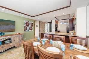 A dining area with a wooden table set for four, a kitchen with modern appliances, and a green accent wall. - Oceanfront 1BD with Panoramic Ocean & Sunset Views -Mahana 406 (Lahaina)