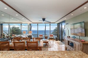 A spacious living area with a large window overlooking the ocean, a ceiling fan, and a television mounted on the wall. - Oceanfront 1BD with Panoramic Ocean & Sunset Views -Mahana 406 (Lahaina)