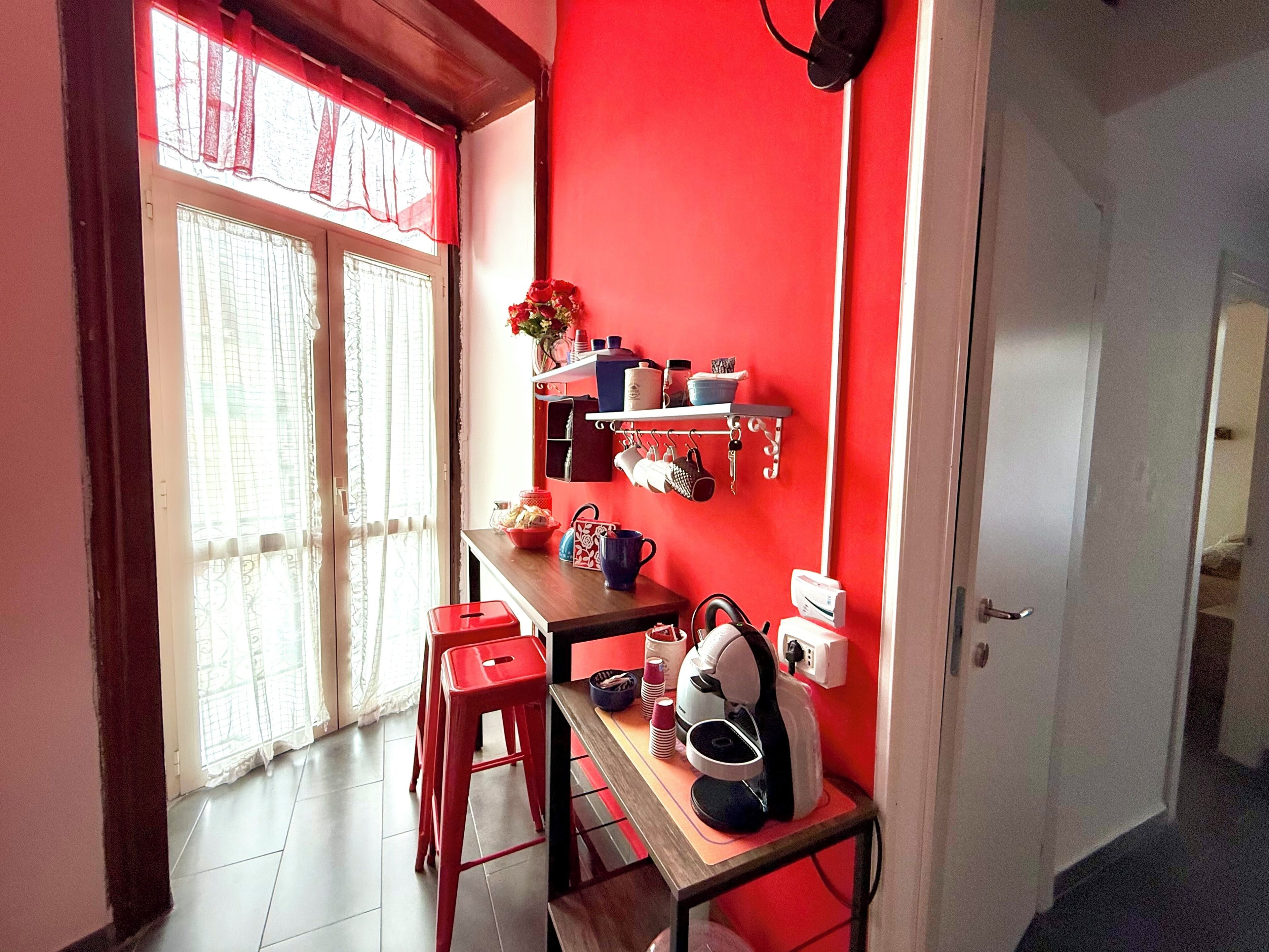 A kitchen with a red wall, a window with curtains, a sink, a countertop with various items, and a door leading to another room.