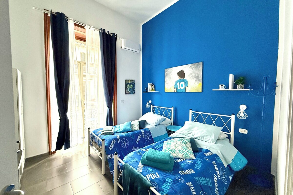 Een slaapkamer met twee bedden, een raam met gordijnen, een blauwe wand en een klein plankje met spullen.