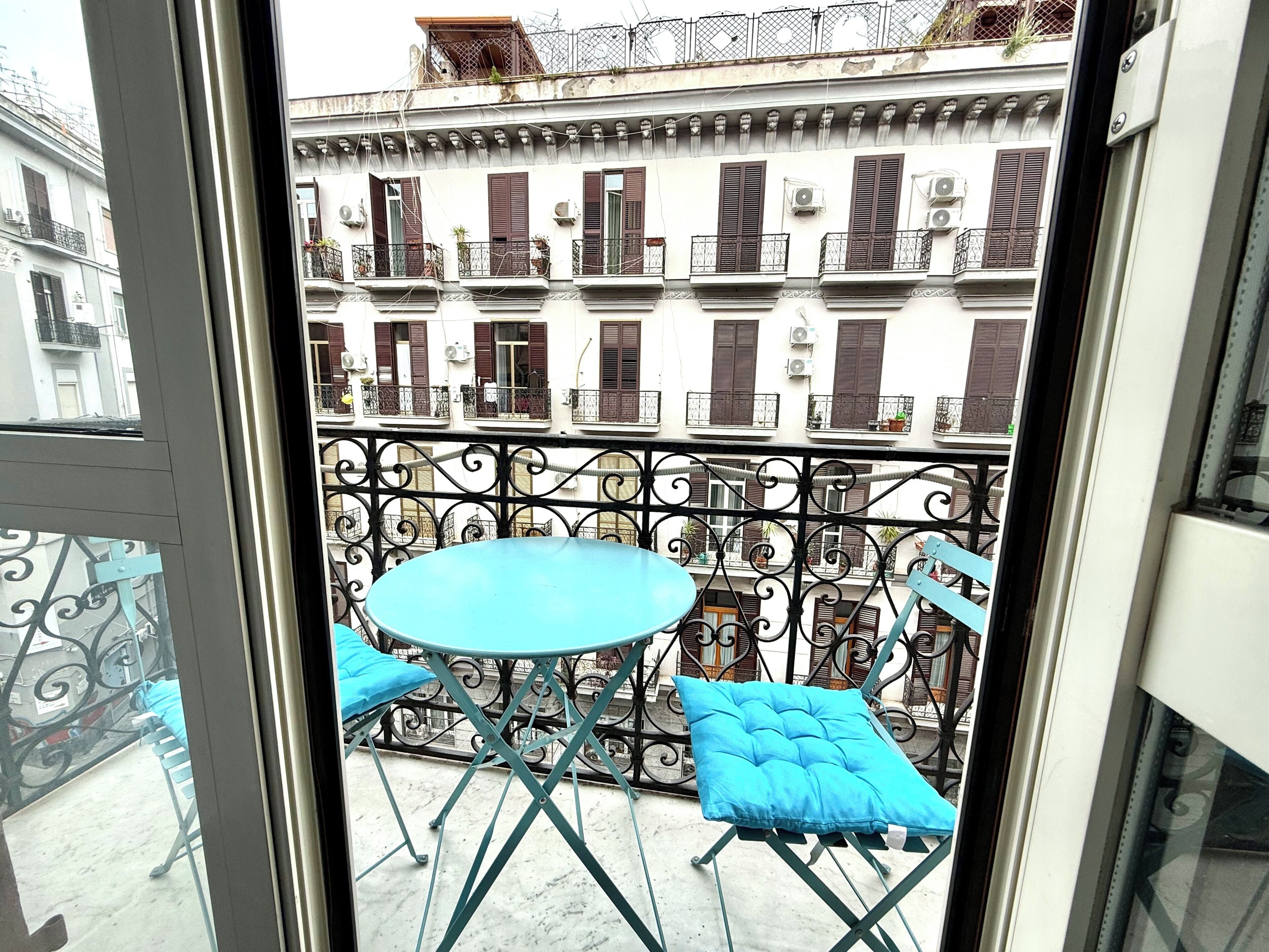 A balcony with a round table and two blue chairs, overlooking a row of buildings with balconies and shutters.