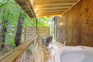 A wooden deck with a hot tub, a table, and chairs, surrounded by trees.