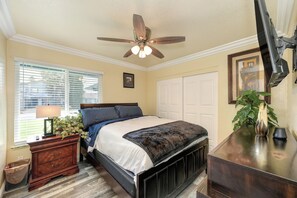 A bedroom with a bed, bedside table, lamp, window with blinds, ceiling fan, and a plant.