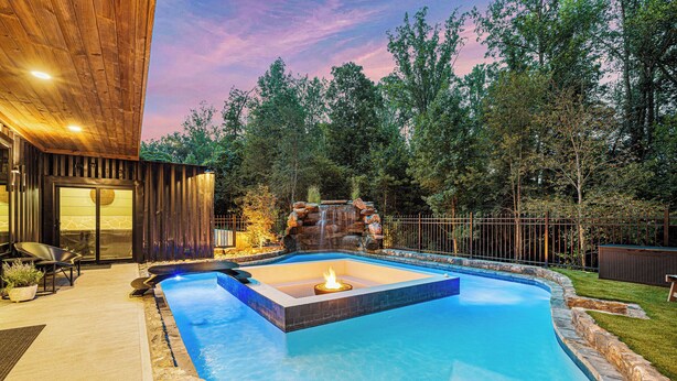 Une piscine avec un foyer, entourée d’une terrasse en pierre et d’une terrasse en bois.