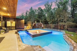 Une piscine avec un foyer, entourée d’une terrasse en pierre et d’une terrasse en bois.